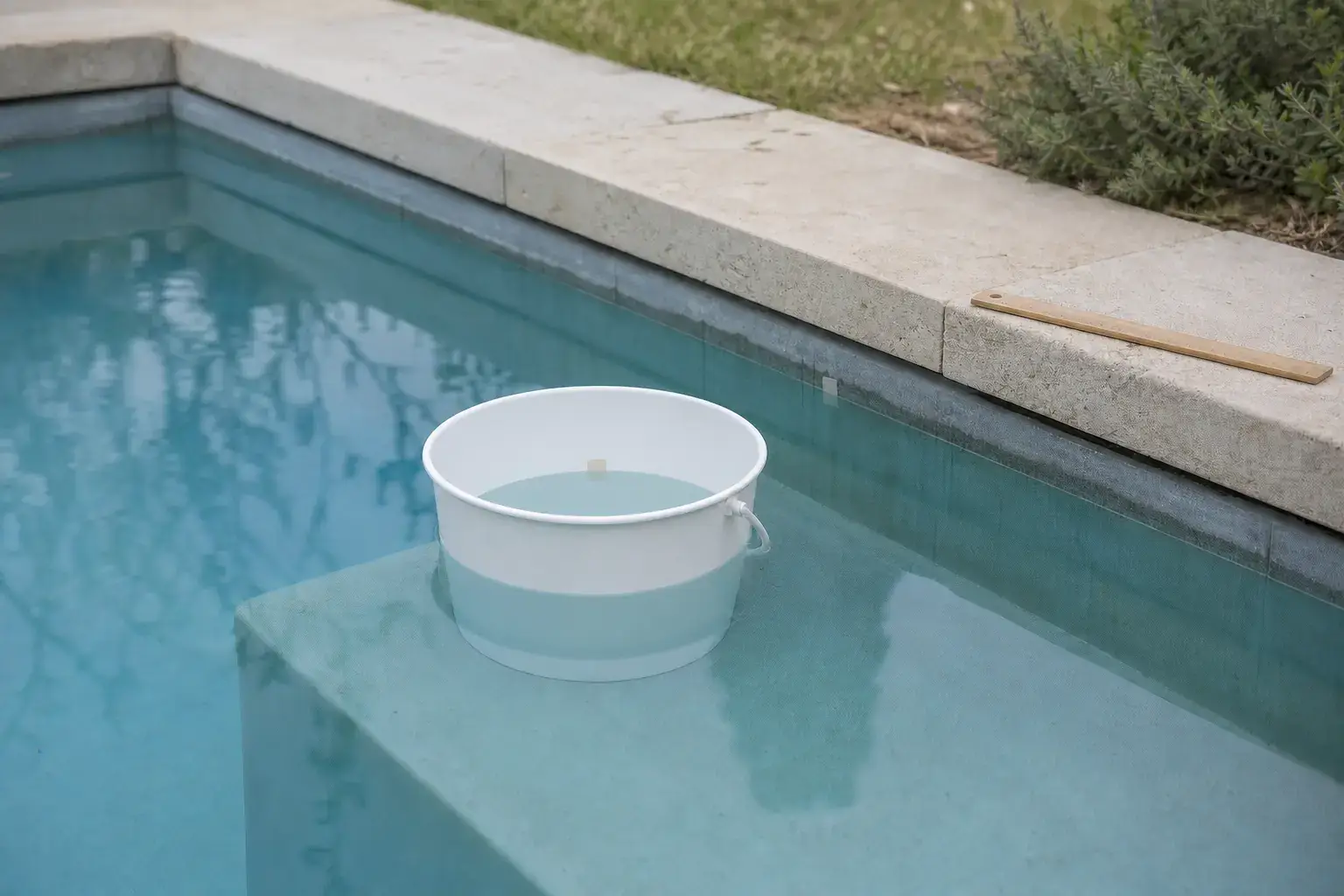 Seau blanc partiellement immergé sur une marche de piscine, avec une règle en bois sur la margelle